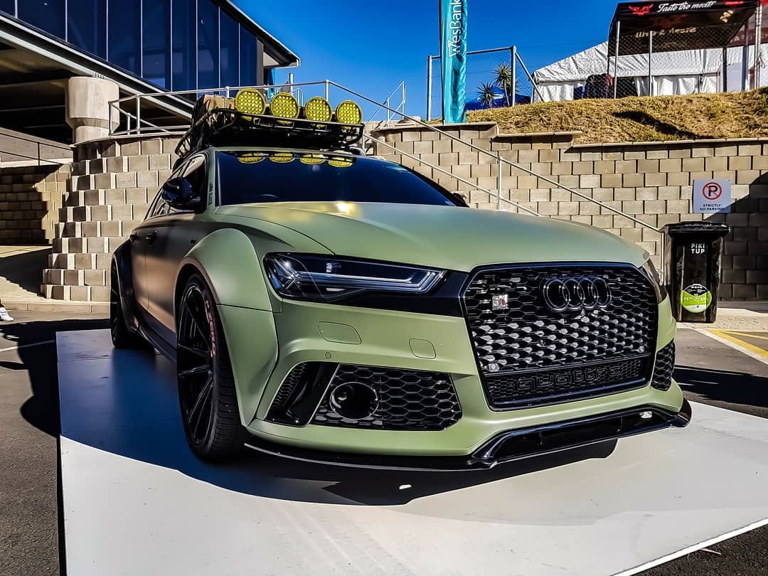 A matt military-green Audi RS6 Avant with roof-mounted lamps, parked at a motoring show.